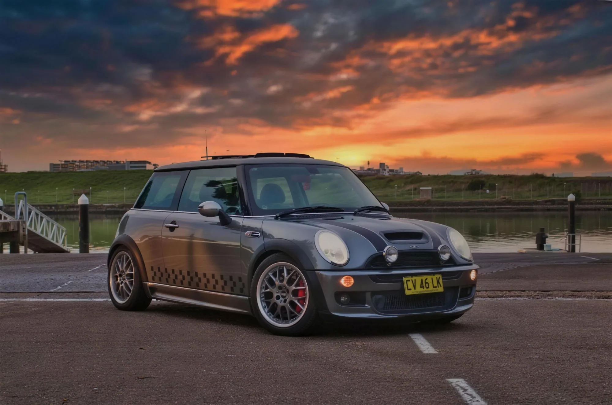 A grey Mini Cooper S parked by the water under an orange sunset sky